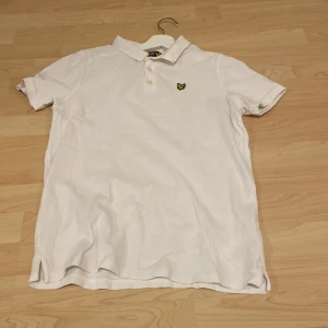 Vit kortärmad pikétröja från Lyle & Scott - Säljer en klassisk vit kortärmad pikétröja från Lyle & Scott med den ikoniska loggan på bröstet. Perfekt för en stilren och avslappnad look. Tröjan har en krage med knappar och är tillverkad i ett bekvämt material. Säljer pga att den är för stor