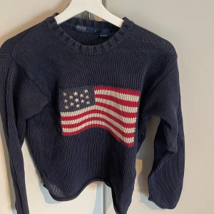Mörkblå stickad tröja med amerikansk flagga från Polo Ralph Lauren - Mörkblå stickad tröja från Polo Ralph Lauren med en stor amerikansk flagga framtill. Tröjan har rund halsringning och långa ärmar. Tillverkad i 100% bomull och har en klassisk, avslappnad passform.