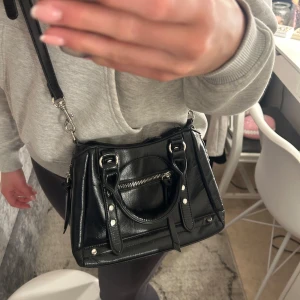 Svart axelväska med dragkedja och nitar - Säljer en svart axelväska med både handtag och justerbar axelrem. Väskan har silverfärgade detaljer, dragkedja framtill och dekorativa nitar. Perfekt storlek för det viktigaste och passar till många olika outfits.