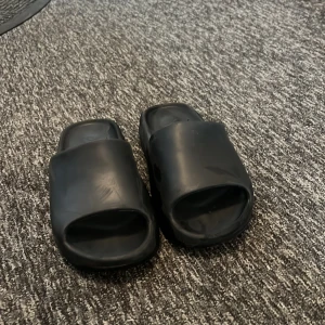 Svarta yeezy slides  - Säljer ett par svarta slides från Adidas. De är tillverkade i syntetmaterial och har en enkel, stilren design med bred rem över foten och räfflad sula för extra grepp. Perfekta att glida i under sommaren eller till och från träningen.