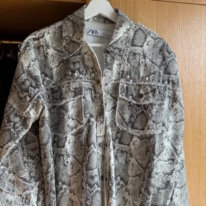 Ormmönstrad jeansjacka med nitar från ZARA - Säljer en grå och svart ormmönstrad jeansjacka från ZARA. Jackan har långa ärmar, två bröstfickor med lock och coola silverfärgade nitar längs överdelen. Perfekt statement-plagg för dig som vill sticka ut.