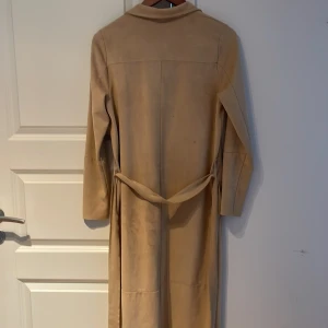 Beige trenchcoat från Gina Tricot - Säljer en beige trenchcoat från Gina Tricot i mockaimitation. Jackan har långa ärmar, två stora fickor framtill och ett bälte i midjan. Perfekt för dig som gillar stilrena och klassiska plagg.