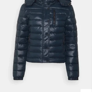 Mörkblå pufferjacka med huva - Säljer en mörkblå pufferjacka. Jackan har huva, dragkedja framtill och två sidofickor med knappar. Den är vadderad och har en glansig yta som ger en modern look. Perfekt för kyligare dagar.