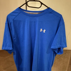 Blå t-shirt från Under Armour - Säljer en blå t-shirt från Under Armour med diskret logga på bröstet. T-shirten är tillverkad i ett syntetmaterial som andas och passar perfekt till träning. Klassisk passform med korta ärmar och rund hals.