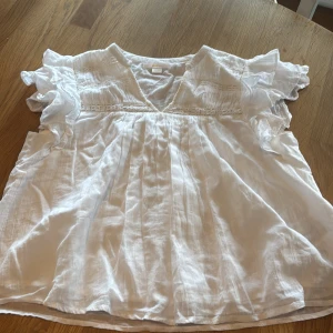 Vit blus med volangärmar från Gina Tricot - Säljer en luftig vit blus från Gina Tricot med v-ringning och fina volangärmar. Blusen har virkade spetsdetaljer och är tillverkad i ett lätt bomullstyg, perfekt för varma dagar.