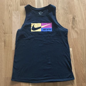 Använd 1 gång Svart linne  från Nike med tryck - Svart linnetopp från Nike med färgglatt tryck framtill och texten 'Just Do It.' Toppen är tillverkad i Dri-Fit material som andas och passar perfekt för träning eller varma dagar.