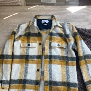 Rutig overshirt från NN07 i ullmix - Säljer en snygg rutig overshirt från NN07 i italienskt tyg. Jackan har stora rutor i beige, grått och gult, två bröstfickor med knappar och klassisk krage. Perfekt lager-på-lager-plagg för höst och vår.