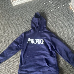 Blå hoodie från Hoodrich - Mörkblå hoodie från Hoodrich med stor vit logga på bröstet. Klassisk huva och magficka framtill. Perfekt för en avslappnad och cool stil.