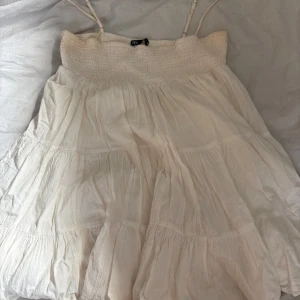 Vit somrig klänning med volanger - Luftig vit klänning med smala axelband och smock upptill. Klänningen har flera volanglager och är tillverkad i ett lätt, skrynkligt tyg . Perfekt för varma dagar och enkel att styla både som klänning eller kjol. Har älskat att bara ta den över bikinin ner till stranden!