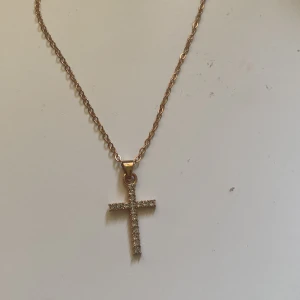 Guldhalsband med korshänge - Elegant guldhalsband med ett glittrande korshänge. Hänget är dekorerat med små stenar som ger en lyxig känsla. Perfekt för att lägga till en touch av elegans till din outfit.