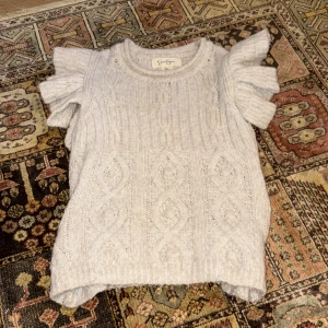 Beige stickad topp med volangärmar från Jessica Simpson - Supersöt beige stickad topp från Jessica Simpson med korta volangärmar och kabelstickat mönster. Perfekt för dig som vill ha något gulligt och unikt i garderoben. Mjuk och skön, passar till många olika stilar.