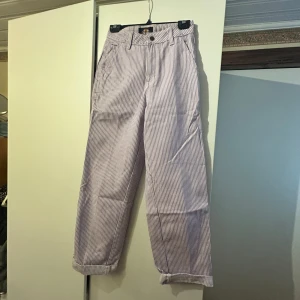Randiga byxor från Dickies - Säljer ett par vita och lila randiga byxor från Dickies med raka ben och normal passform med skickar stil. Byxorna har fickor både fram och bak samt klassisk Dickies-logga på bakfickan. 
