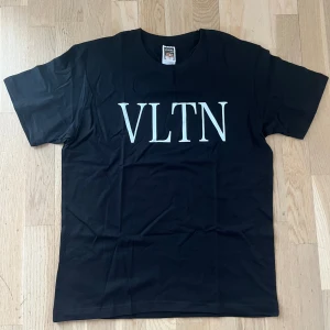 Svart t-shirt med VLTN-tryck - Snygg svart t-shirt med stort vitt VLTN-tryck på bröstet. Klassisk passform och rund halsringning. Perfekt för en stilren och trendig look.
