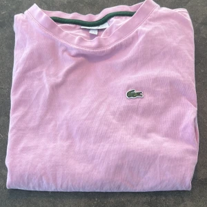 Ljusrosa t-shirt från Lacoste - Säljer en ljusrosa t-shirt från Lacoste med klassisk krokodillogga broderad på bröstet. T-shirten har rund halsringning med grön detalj på insidan och är tillverkad i mjuk bomull. Perfekt för en avslappnad och stilren look.