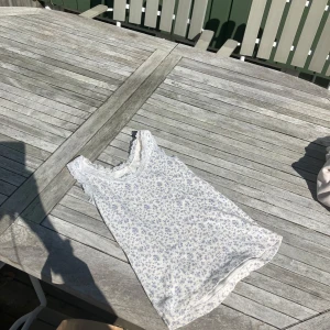 Vit blommig linnetopp med spets från Brandy Melville - Supersöt vit linnetopp från Brandy Melville med små blå blommor och spetsdetaljer vid halsringning och axlar. Tillverkad i mjuk bomull, perfekt för varma dagar. Säljes då den inte kommer till användning längre! Använd fåtal gånger och köpte den för 250kr och den säljs inte längre! 