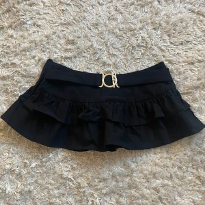 Svart volangkjol med bälte - Säljer svart kortkjol med volang och brett midjebälte! Guldfärgat spänne och en dragkedja i sidan. Super snygg till fester!