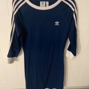 klänning  - Sportig tight blå klänning från Adidas med klassiska vita ränder längs axlar och ärmar samt vit ribbad halsringning och ärmslut. Klänningen har trekvartsärm och en liten Adidas-logga på bröstet. Pris kan diskuteras vid snabb affär