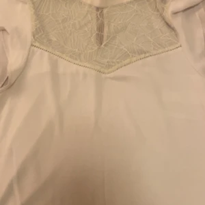 Beige topp med spetsdetaljer - Elegant beige topp med vackra spetsdetaljer vid halsringningen. Topp har en stilren design med en liten öppning i ryggen som ger en extra touch. Perfekt för speciella tillfällen.