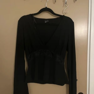 Svart långärmad topp med spets från Gina Tricot - Snygg svart långärmad topp från Gina Tricot med v-ringning och spetsdetaljer vid midjan. Toppen har en figurnära passform och är perfekt för att lyfta din outfit med en elegant touch.