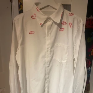 Vit skjorta med läppstiftsmönster från Maison Margiela - Vit skjorta från Maison Margiela med unikt läppstiftsmönster i rött över krage och axlar. Klassisk krage, bröstficka och knäppning framtill. Perfekt för dig som vill sticka ut med en cool detalj. obs one to one