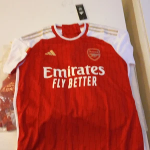 Arsenal hemmatröja 2023/24 från Adidas - Säljer en Arsenal hemmatröja från Adidas i rött med vita ärmar och guldiga detaljer. Tröjan har klubbmärke och sponsortryck på bröstet samt klassiskt mönster. Perfekt för dig som vill visa ditt stöd för Arsenal på matchdagar!
