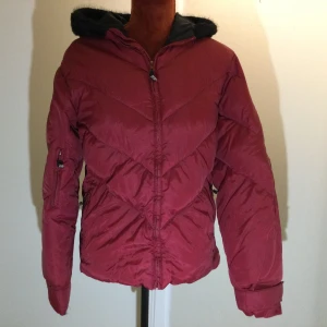Dunjacka Röd strl: M - Säljer en vinröd pufferjacka med huva och svart foder. Jackan har dragkedja framtill, långa ärmar och en sportig look. Perfekt för kyliga dagar och har en klassisk passform. Snygg och enkel att matcha till olika outfits. Välanvänd med smådefekter, se bilder! Dragkedja 2 framfickor, 1 innerficka Huva med fuskpäls
