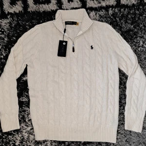 Vit stickad tröja med dragkedja från Polo Ralph Lauren - Säljer en klassisk vit stickad tröja från Polo Ralph Lauren med halv dragkedja och kabelstickat mönster. Tröjan har lång ärm, ribbade muddar och den ikoniska broderade loggan på bröstet. Perfekt för lager-på-lager-stil.(malmö)