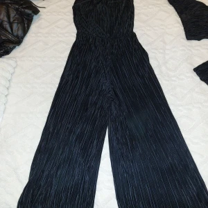 Svart byxdress med smala axelband - Säljer en svart byxdress med smala axelband och lätt plisserat tyg. Byxdressen har vida ben och resår i midjan för en bekväm passform. Perfekt för dig som vill ha en stilren och trendig look.