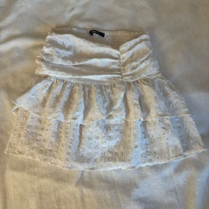 Vit volangkjol från Gina Tricot - Supersöt vit kjol från Gina Tricot med volanger och smock i midjan. Kjolen har ett diskret rutigt mönster i tyget och är kort i modellen. Perfekt för sommarens alla tillfällen!