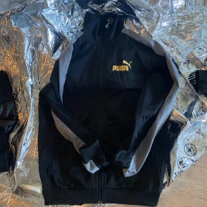 Svart träningsjacka från Puma - Svart träningsjacka från Puma med dragkedja och gul logga på bröstet. Jackan har långa ärmar med gråa och vita ränder längs sidorna. Perfekt för sportiga tillfällen.