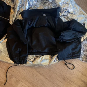 Svart croppad jacka med dragsko - Säljer en svart croppad jacka med dragsko i midjan och vid ärmsluten. Jackan har långa ärmar och en hög krage. Perfekt för dig som vill ha en stilren och enkel look.