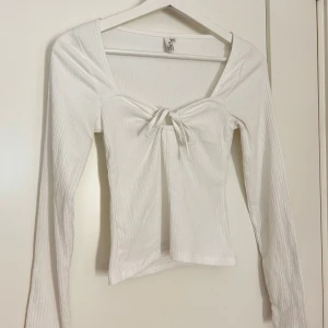 Vit långärmad topp med knyt - Säljer en vit långärmad topp med fyrkantig ringning och knyt framtill. Toppen är ribbad och har en feminin passform som passar perfekt till jeans eller kjol.
