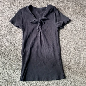 Svart ribbad kortärmad med knyt från The Black Label Collection - Svart ribbad tröja i 100% bomull från The Black Label Collection (lager 157) Kortärmad modell med v-ringning och dekorativ knytning framtill. Enkel och stilren design som passar perfekt till sommaren. På en utav bilderna såg tröjan grå ut men tröjan är svart. Materialet är stretchig.