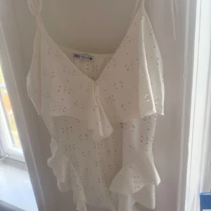 Vit volangig linnetopp från Zara - Säljer en söt vit linnetopp från Zara med broderade hålmönster och volangdetaljer. Toppen har tunna axelband som knyts och en luftig, flowy passform. Perfekt för varma dagar och somriga outfits!