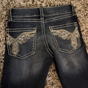 Mörkblå bootcut jeans med broderade bakfickor - Snygga mörkblå jeans med bootcut passform och broderade detaljer med paljetter på bakfickorna. Jeansen har kontrastsömmar och klassisk femficksdesign. Perfekta för dig som gillar lite extra detaljer på dina jeans.
