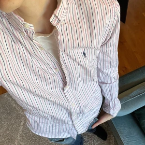 Stilren Randig skjorta från Polo Ralph Lauren - Säljer en klassisk vit skjorta med rosa och marinblå ränder från Polo Ralph Lauren. Skjortan har lång ärm, knappar framtill och broderad logga på bröstet. Slim fit-modell som passar perfekt till många tillfällen.