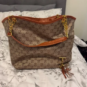 Beige och brun axelväska med kedjedetaljer - Säljer en beige och brun axelväska med monogrammönster och guldfärgade kedjor. Väskan har bruna detaljer i syntetmaterial, tofs och innerfack med dragkedja. Perfekt för dig som vill ha en rymlig och snygg accessoar.