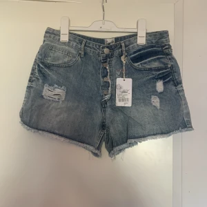 Blå jeansshorts med slitningar och knappar - Snygga blå jeansshorts med slitna detaljer och råa kanter. Modellen har hög midja och knäppning framtill med flera knappar. Perfekta för sommaren och ger en cool, avslappnad look.