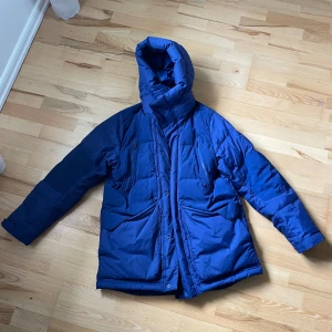 Blå Nike dunjacka med huva - Säljer en blå dunjacka med huva och flera praktiska fickor. Jackan har lång ärm och dragkedja framtill, perfekt för kalla dagar. Passformen är normal och jackan är vadderad för extra värme.