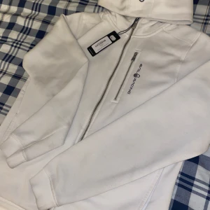 Vit hoodie med dragkedja från Sail Racing - Vit hoodie från Sail Racing med dragkedja och huva. Snyggt broderad logga på bröstet och en bröstficka med dragkedja. Perfekt för en sportig och stilren look.