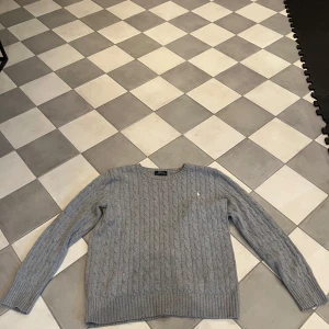 Grå stickad tröja från Polo Ralph Lauren - Säljer en klassisk grå stickad tröja från Polo Ralph Lauren. Tröjan har rund halsringning och ett diskret broderat logomärke på bröstet. Perfekt för en stilren och avslappnad look.