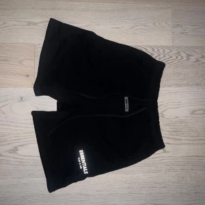 Svarta shorts från Essentials Fear of God - Svarta shorts från Essentials Fear of God med vit logga på vänster ben. Modellen har normal passform, resår i midjan och snörning framtill. Tillverkade i mjukt bomullsmaterial, perfekta för en avslappnad stil.