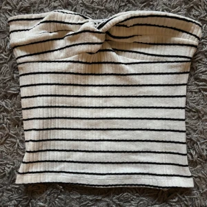 Randig tubtopp från NA-KD - Säljer en vit tubtopp med svarta ränder från NA-KD. Toppen är ribbad och har en twistad detalj framtill. Perfekt för sommaren och enkel att styla med jeans eller kjol💕🫶🏼