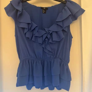 Blå volangtopp från H&M - Säljer en blå topp från H&M med volangdetaljer längs ringningen och nederkanten. Toppen är ärmlös och har Resår i midjan. Skulle säga att den passar mellan xl-s beror hur man vill ha passformen. Slutsåld överallt💞