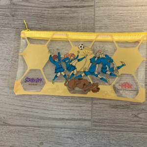 Transparent gul Scooby-Doo pen fodral  - Säljer en transparent gul pen fodral med Scooby-Doo-tryck där karaktärerna spelar fotboll. Väskan har dragkedja upptill och hexagonmönster i plast. Perfekt för att hålla småsaker organiserade.