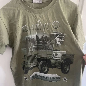 Olivgrön t-shirt med army-tryck från Survivors - Olivgrön t-shirt från Survivors med stort army-tryck både fram och bak. Motiv med militärjeep, soldater och texten 'ARMY' samt 'THE UNSTOPPABLE LEGEND'. Rund halsringning och normal passform. 100% bomull. Och det passar super för dig som vill ha en Y2K känsla i din garderob!