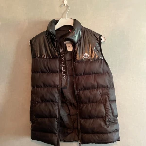 Svart dunväst från Moncler - Säljer en svart dunväst från Moncler med glansig ovandel och klassisk Moncler-logga på bröstet. Västen har hög krage och dragkedja framtill. Perfekt för lager-på-lager och stilren look.