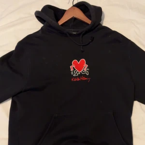 Svart hoodie från Axel Arigato x Keith Haring - Svart hoodie från Axel Arigato med Keith Haring-tryck på bröstet i rött och vitt. Klassisk huva med dragsko och känguruficka framtill. Mjuk bomullskvalitet och relaxed fit. 