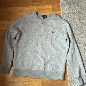 Grå sweatshirt från Polo Ralph Lauren Junior - Säljer en stilren grå sweatshirt från Polo Ralph Lauren med klassisk rund halsringning och broderad logga på bröstet. Tröjan har långa ärmar och ribbade muddar vid ärmslut och nederkant. Perfekt för en avslappnad look.