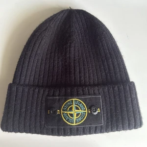  Stone Island mössa - Svart ribbstickad mössa från Stone Island med uppvikt kant och klassisk logotyp framtill. Märkeslapp på sidan och insida. Perfekt för kyliga dagar och stilren streetwear-look.
